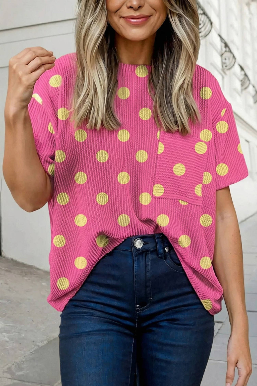 A person in the Aapparella Texture Polka Dot Chest Pocket T Shirt, a pink ribbed short-sleeve top, tucked into high-waisted dark jeans, stands on a sidewalk.