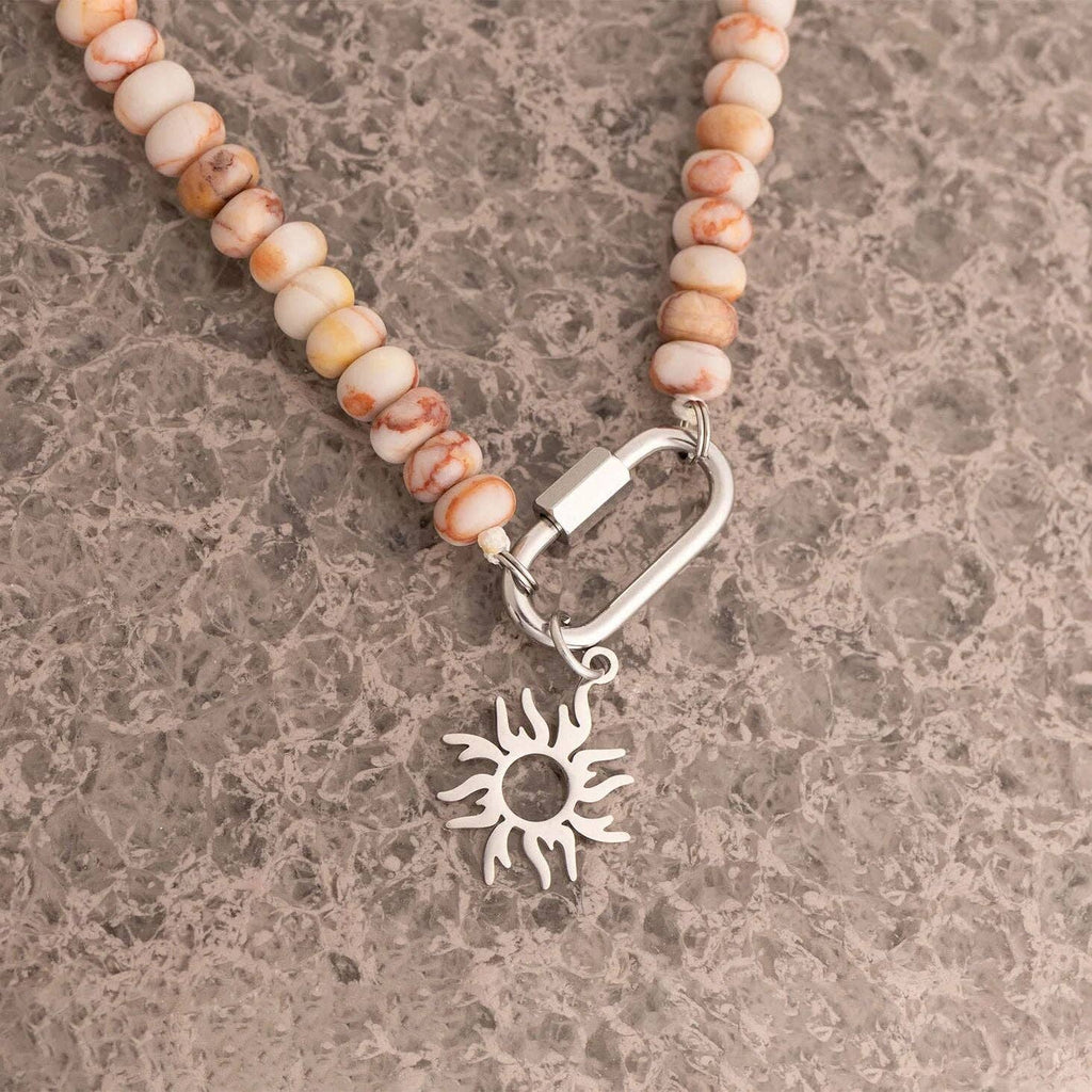 WJW Stainless Steel Chunky Glass Bead Carabiner Necklace featuring beige and brown stones, a silver-tone carabiner clasp, and a metallic sun-shaped pendant, displayed on a textured gray surface.