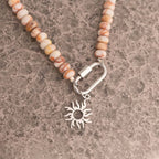 WJW Stainless Steel Chunky Glass Bead Carabiner Necklace featuring beige and brown stones, a silver-tone carabiner clasp, and a metallic sun-shaped pendant, displayed on a textured gray surface.