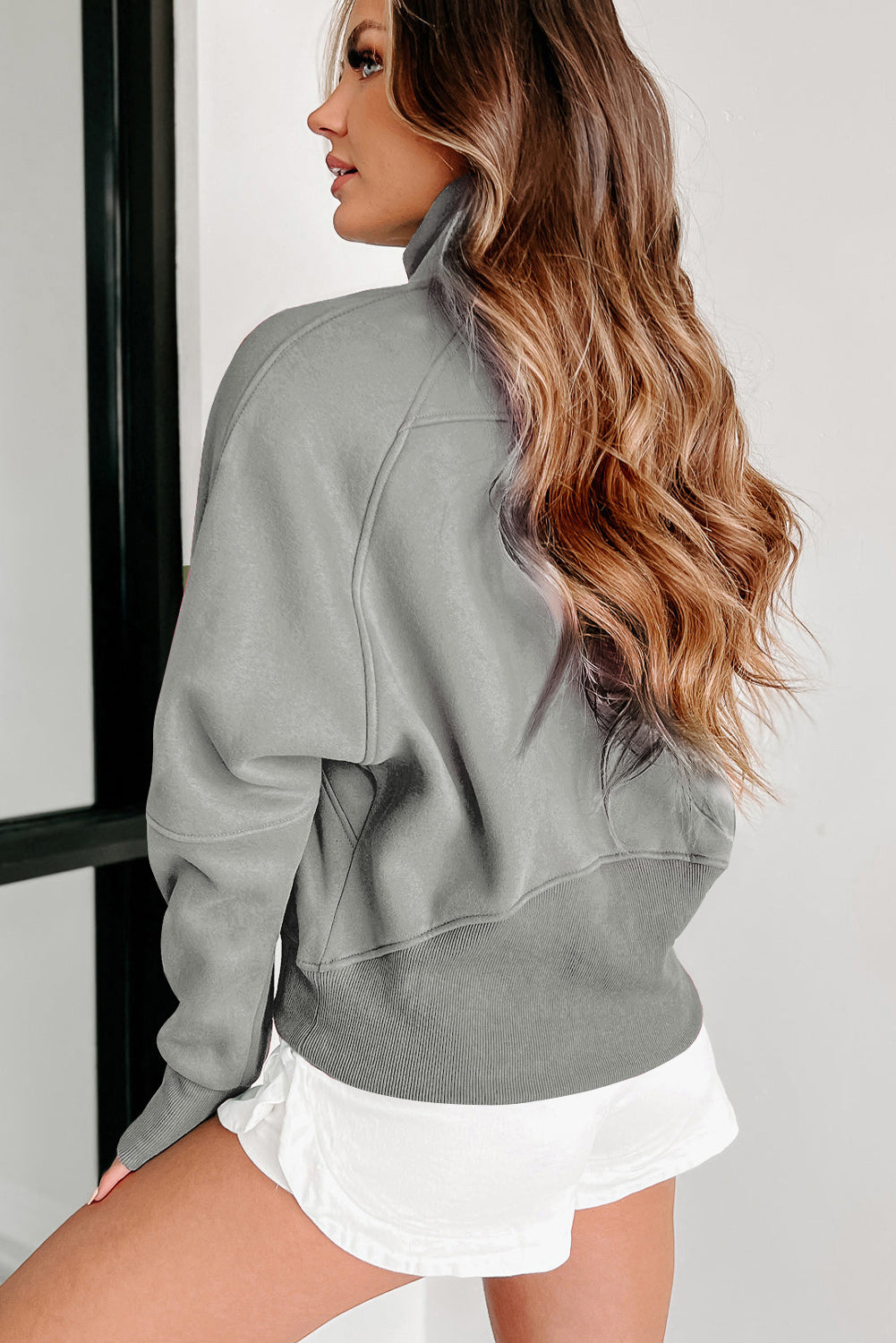 A woman with long wavy hair wears a Shewin gray zip-up stand collar ribbed thumbhole sleeve sweatshirt made of high-quality material and white shorts.