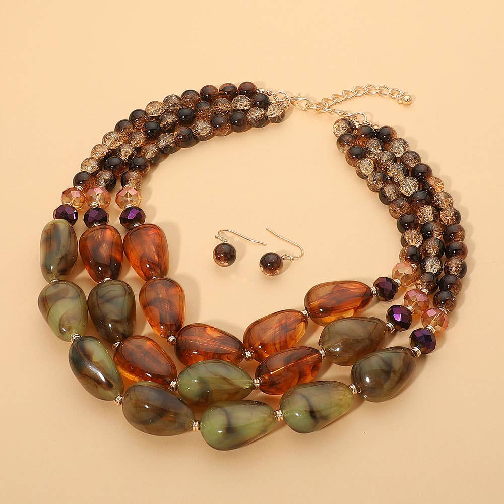 Introducing the Chunky Beaded Layered Statement Necklace by Sensibling Corp.—a bold three-strand necklace set designed with green and brown oversized beads, complemented by matching round bead drop earrings. Elegantly showcased on a beige background, this set is perfect for those who appreciate standout jewelry pieces.