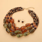 Introducing the Chunky Beaded Layered Statement Necklace by Sensibling Corp.—a bold three-strand necklace set designed with green and brown oversized beads, complemented by matching round bead drop earrings. Elegantly showcased on a beige background, this set is perfect for those who appreciate standout jewelry pieces.