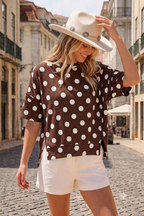 A woman wearing the Celeste Clothing Women’s Polka Dot Rib Knit Top, styled with a beige hat and white shorts, stands on a cobblestone city street, looking down and smiling.