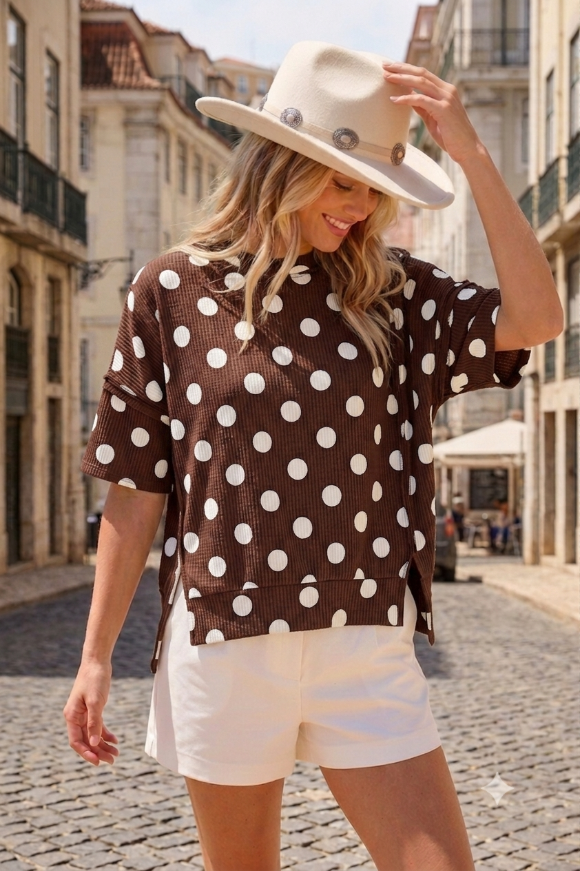 A woman wearing the Celeste Clothing Women’s Polka Dot Rib Knit Top, styled with a beige hat and white shorts, stands on a cobblestone city street, looking down and smiling.