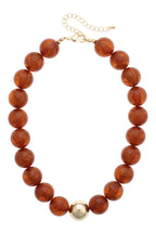 The Marjorie Fall Beaded Necklace by Canvas Style features oversized amber-colored resin beads accented with a single large gold bead at the center and is finished with an adjustable gold clasp.