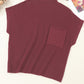 The Shewin Patch Pocket Ribbed Knit Short Sleeve Sweater in burgundy, ideal for transitional weather, is displayed on a surface alongside a magazine and glasses. This comfortable sweater includes a convenient front pocket for small essentials.