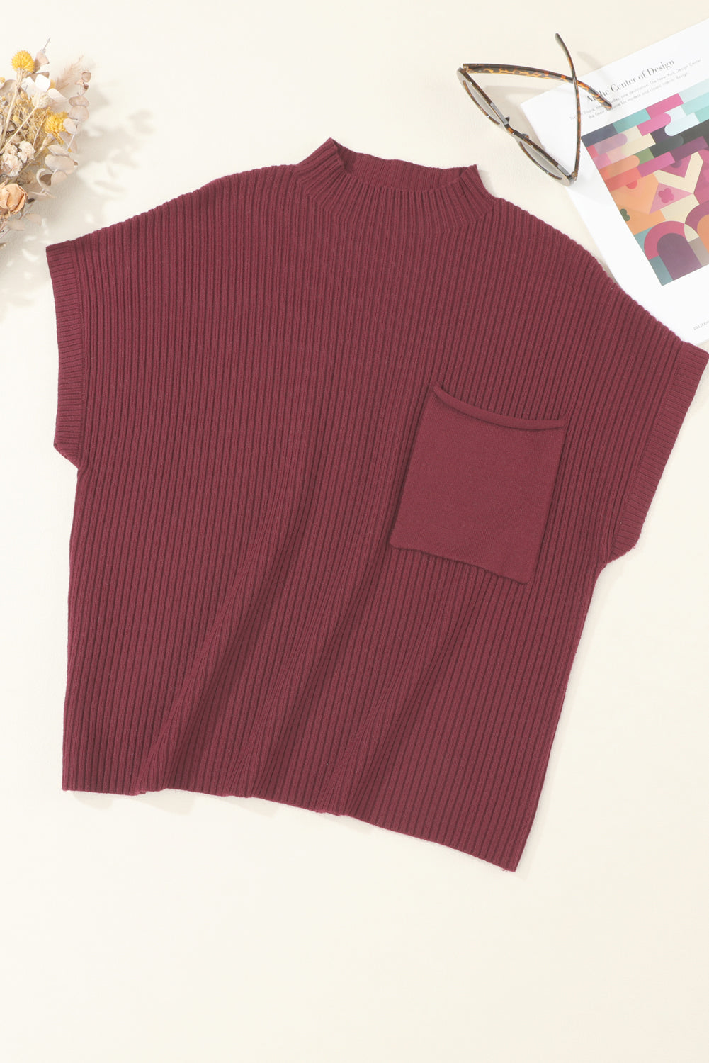 The Shewin Patch Pocket Ribbed Knit Short Sleeve Sweater in burgundy, ideal for transitional weather, is displayed on a surface alongside a magazine and glasses. This comfortable sweater includes a convenient front pocket for small essentials.