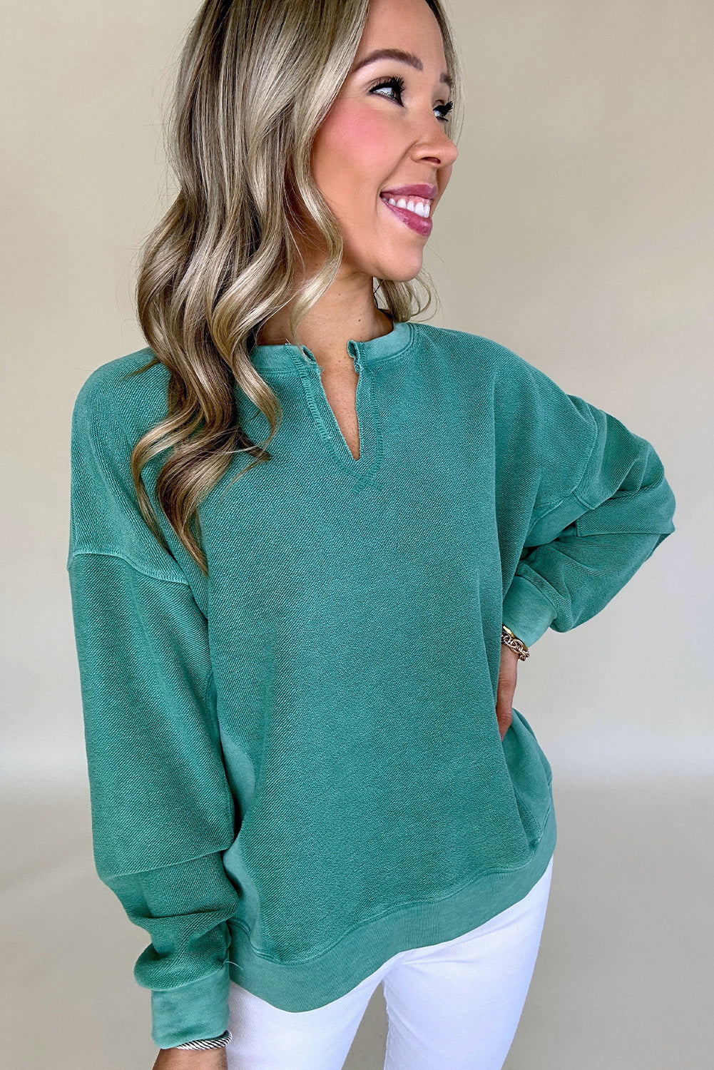 A person with long hair smiles, wearing a Notched Neck Exposed Seam Drop Shoulder Sweatshirt by Shewin and white pants, standing against a plain background.