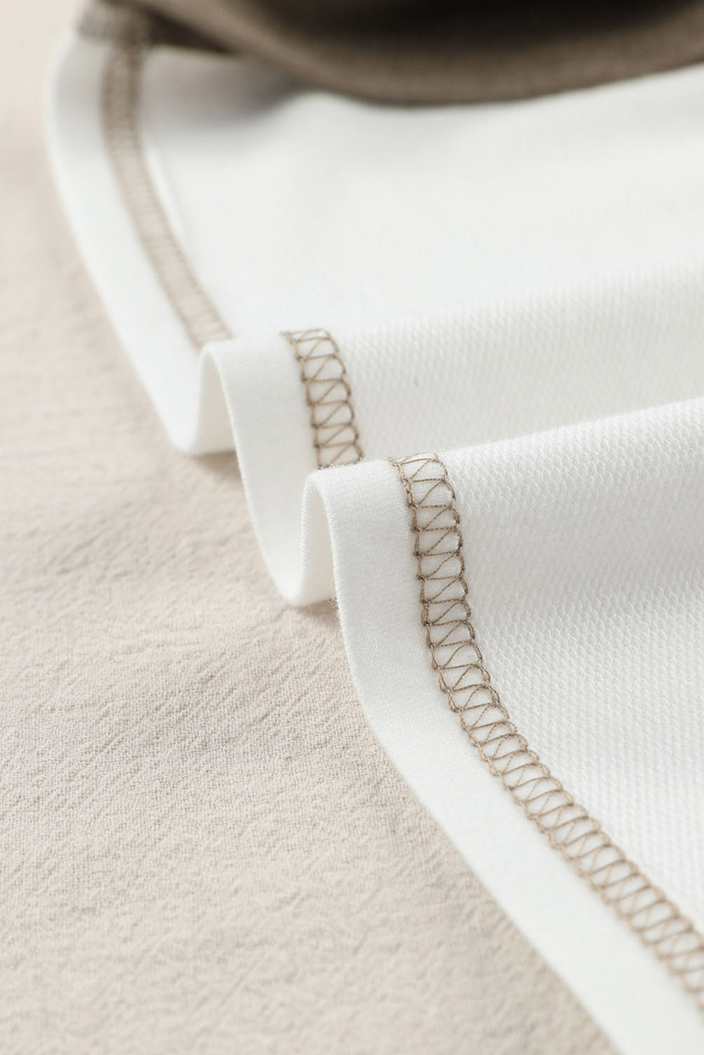 Close-up of the Shewin Khaki Color Block Exposed Seam Long Sleeve Top, showcasing its brown stitching along the edge and unique patchwork design, placed on a beige surface.