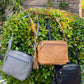 Three Roma Leathers, Inc. Cowhide Leather Crossbody bags in gray, tan, and black, each with adjustable shoulder straps, hang on a green bush against a brick wall backdrop.