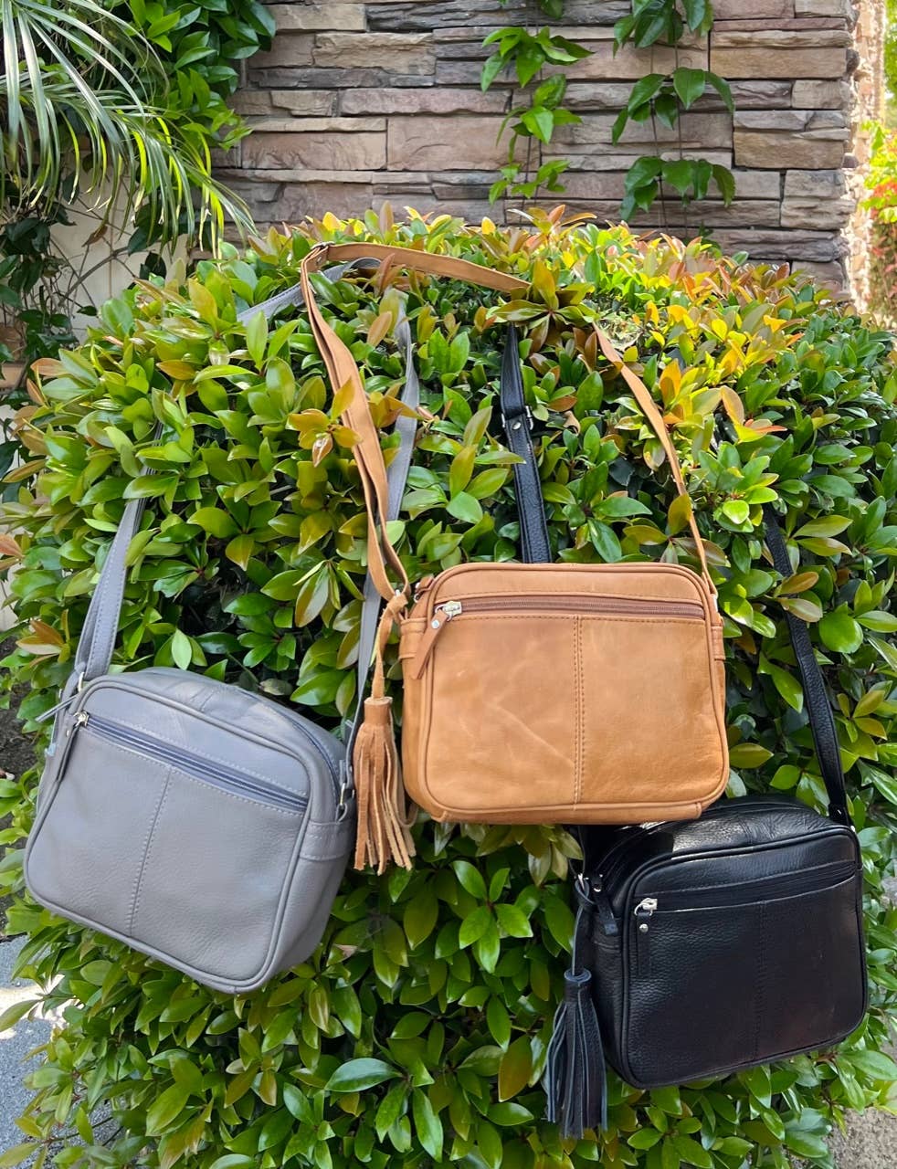 Three Roma Leathers, Inc. Cowhide Leather Crossbody bags in gray, tan, and black, each with adjustable shoulder straps, hang on a green bush against a brick wall backdrop.