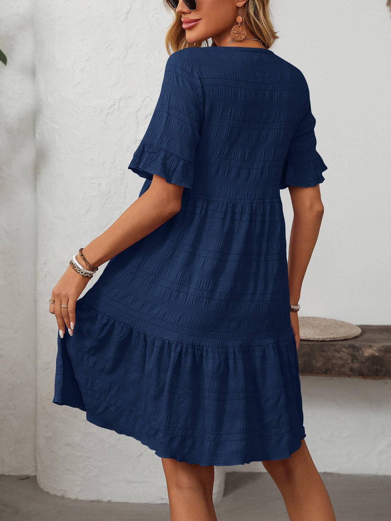 A woman models the Suhkasana Loose Casual Short-Sleeve Dress in navy blue, featuring a textured finish and ruffled hem, seen indoors from the back.