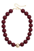 The Marjorie Game Day Beaded Necklace from Canvas Style features oversized burgundy beads accented by a single central gold bead, finished with a gold chain and clasp for adjustable length—a bold statement necklace designed to elevate any outfit.