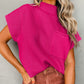 Wearing the Shewin Patch Pocket Ribbed Knit Short Sleeve Sweater in bright pink, paired with brown pants, a person stands against a textured gray wall. Their wavy, light-colored hair reaches their shoulders, making this top ideal for transitional weather.