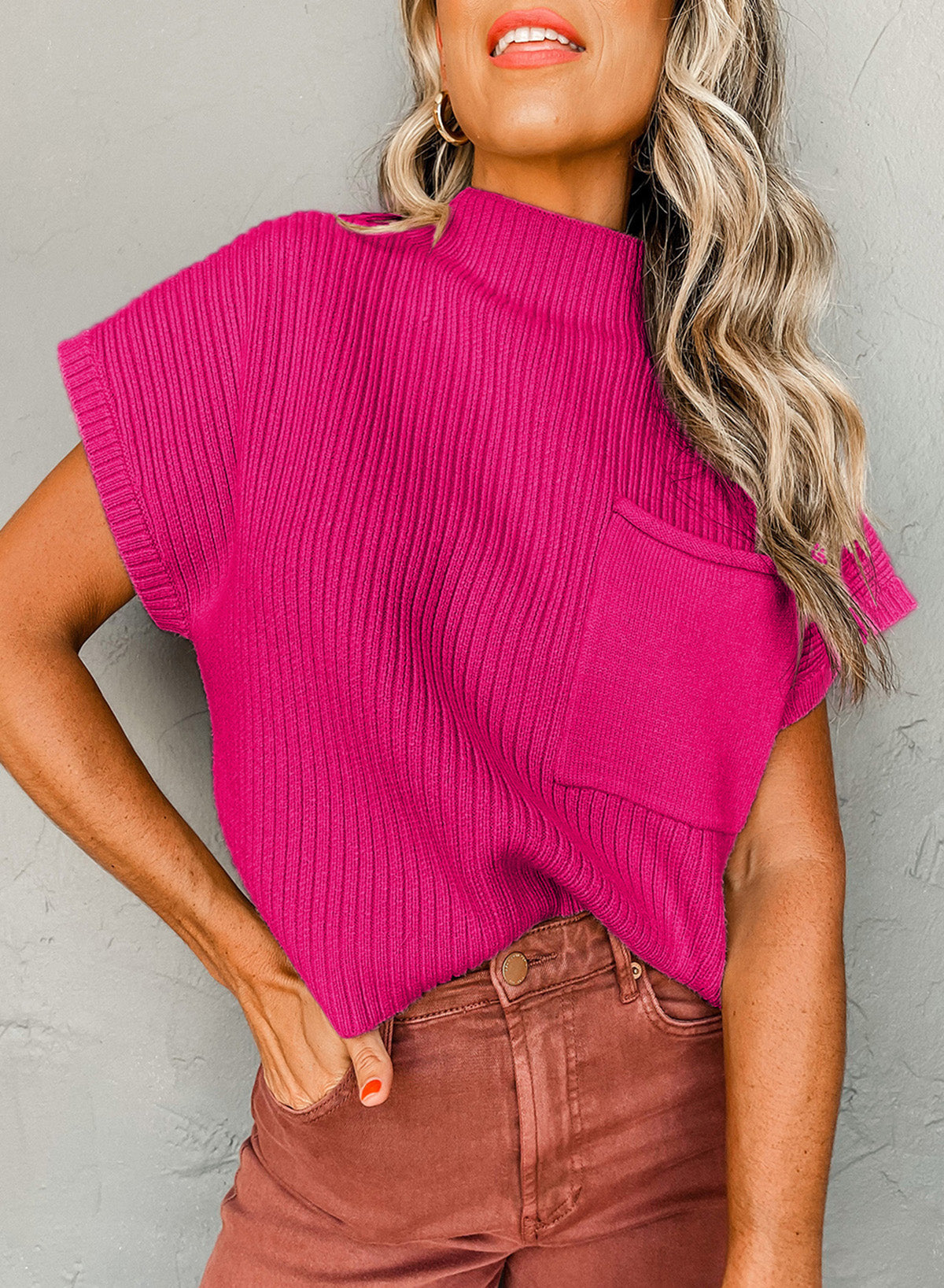 Wearing the Shewin Patch Pocket Ribbed Knit Short Sleeve Sweater in bright pink, paired with brown pants, a person stands against a textured gray wall. Their wavy, light-colored hair reaches their shoulders, making this top ideal for transitional weather.