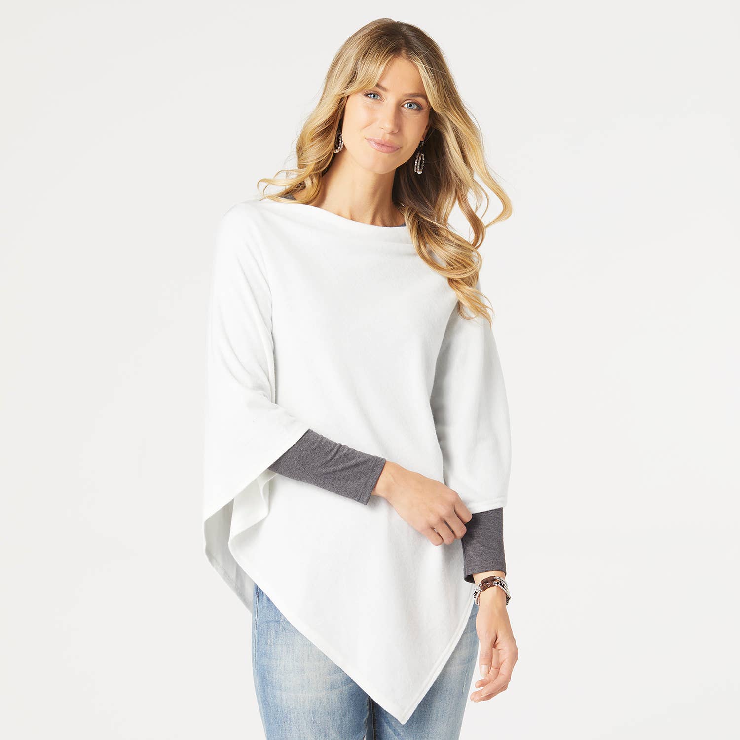 A woman with long blonde hair wears the COCO + CARMEN Lightweight Brushed Poncho Wrap over a gray long-sleeve shirt and blue jeans, standing against a plain white background.