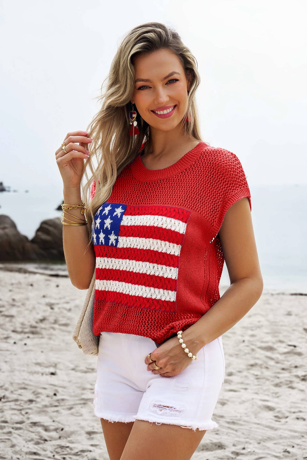 A woman smiles at the camera on a beach, wearing 1 the Woman’s Flag Graphic Short Sleeve Sweater Top with white shorts. Rocks and ocean in the background highlight her chic, patriotic look.