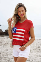 A woman smiles at the camera on a beach, wearing 1 the Woman’s Flag Graphic Short Sleeve Sweater Top with white shorts. Rocks and ocean in the background highlight her chic, patriotic look.