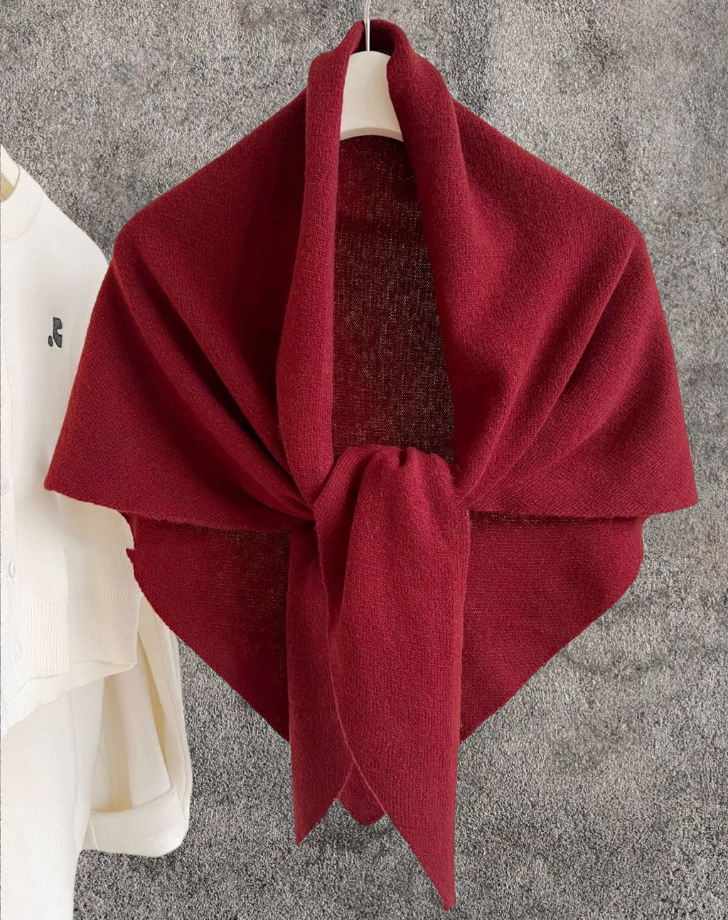 A JC Sunny Fashion Cashmere-Blend Knit Triangle Shawl with a cross-front design in deep red is draped over a hanger against a gray textured wall, next to a white shirt hanging on the left.