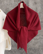 A JC Sunny Fashion Cashmere-Blend Knit Triangle Shawl with a cross-front design in deep red is draped over a hanger against a gray textured wall, next to a white shirt hanging on the left.