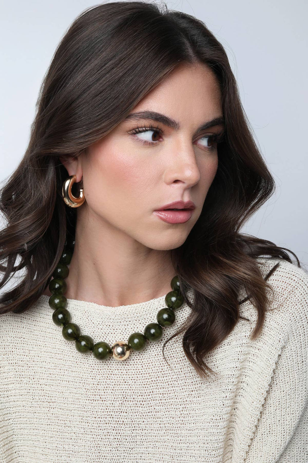 A woman with wavy brown hair is wearing a cream knit sweater, gold hoop earrings, and the Marjorie Fall Beaded Necklace by Canvas Style, featuring chunky green resin beads, as she looks to her left against a plain background.