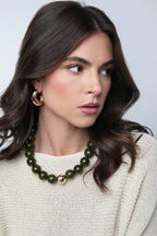 A woman with wavy brown hair is wearing a cream knit sweater, gold hoop earrings, and the Marjorie Fall Beaded Necklace by Canvas Style, featuring chunky green resin beads, as she looks to her left against a plain background.