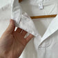 A hand holds the collar of a Rosa Clothing White Relaxed Short Sleeve Blouse on a wooden hanger, showing the size small tag inside the neckline.