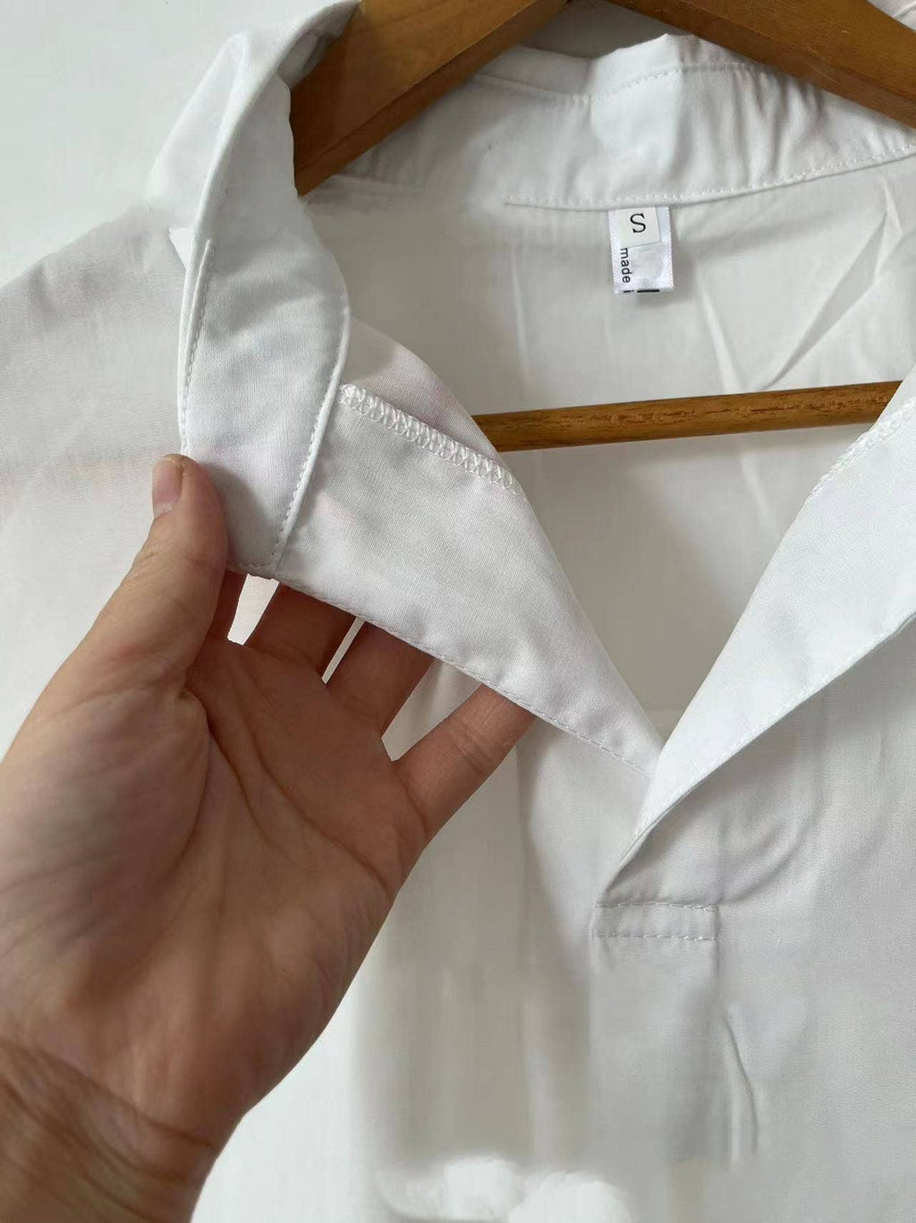 A hand lifts the collar of the FASHION GO White Relaxed Fit Short Sleeve Blouse on a wooden hanger, revealing the inner stitching of the collar and a visible size S tag.