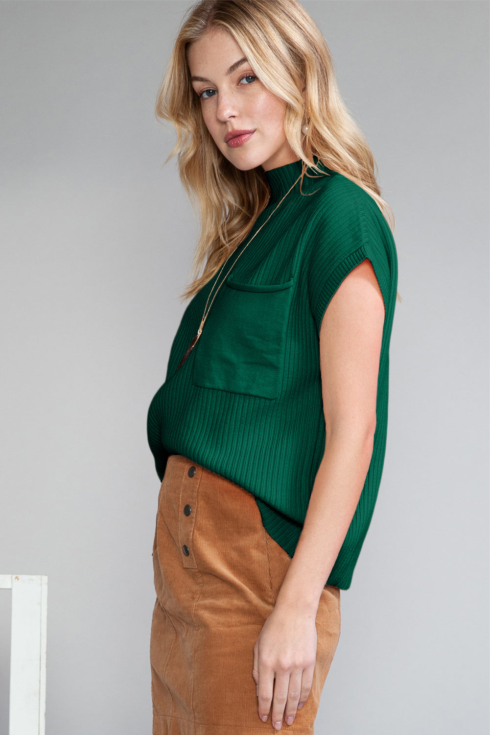 A woman with long blonde hair stands against a plain backdrop, wearing Shewin's Patch Pocket Ribbed Knit Short Sleeve Sweater in green and a brown skirt, offering versatile style perfect for changing weather.