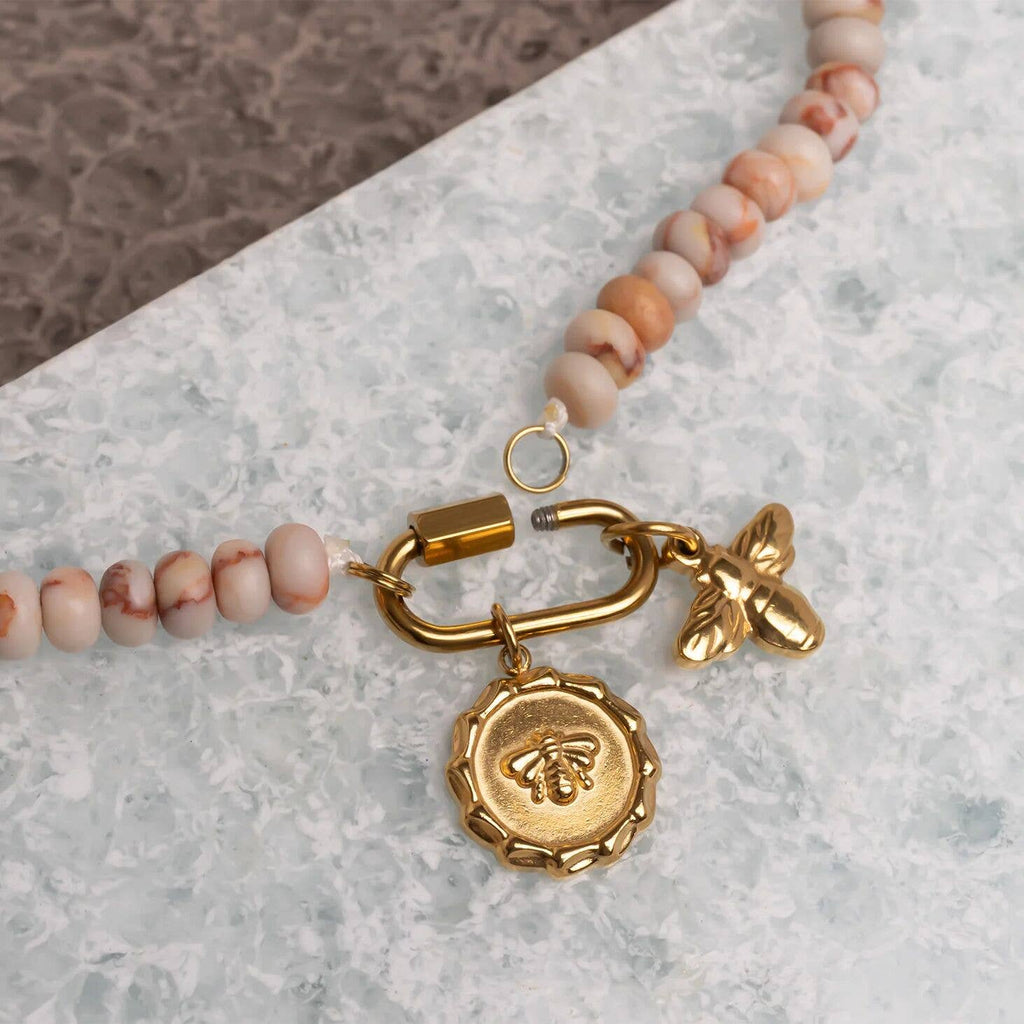 The WJW Stainless Steel Chunky Glass Bead Carabiner Necklace features a gold-toned clasp, accented with a round bee-design charm and a small gold bee charm, displayed on a textured surface.