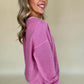 A woman smiles while wearing the Shewin Notched Neck Exposed Seam Drop Shoulder Sweatshirt in pink paired with white pants, set against a plain background.