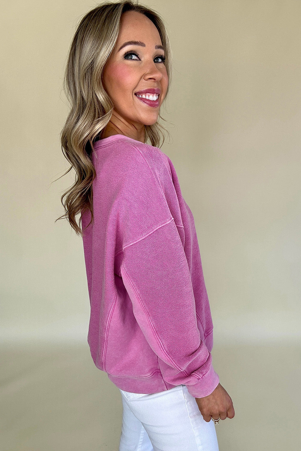 A woman smiles while wearing the Shewin Notched Neck Exposed Seam Drop Shoulder Sweatshirt in pink paired with white pants, set against a plain background.