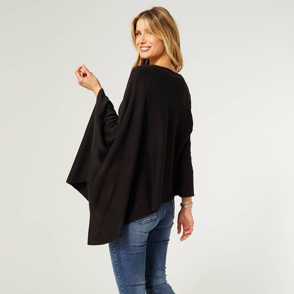 A woman stands smiling in COCO + CARMEN's Lightweight Ponchos Wrap, styled with blue jeans and posing with her back slightly turned—a chic, versatile piece for effortless layering.