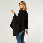 A woman stands smiling in COCO + CARMEN's Lightweight Ponchos Wrap, styled with blue jeans and posing with her back slightly turned—a chic, versatile piece for effortless layering.