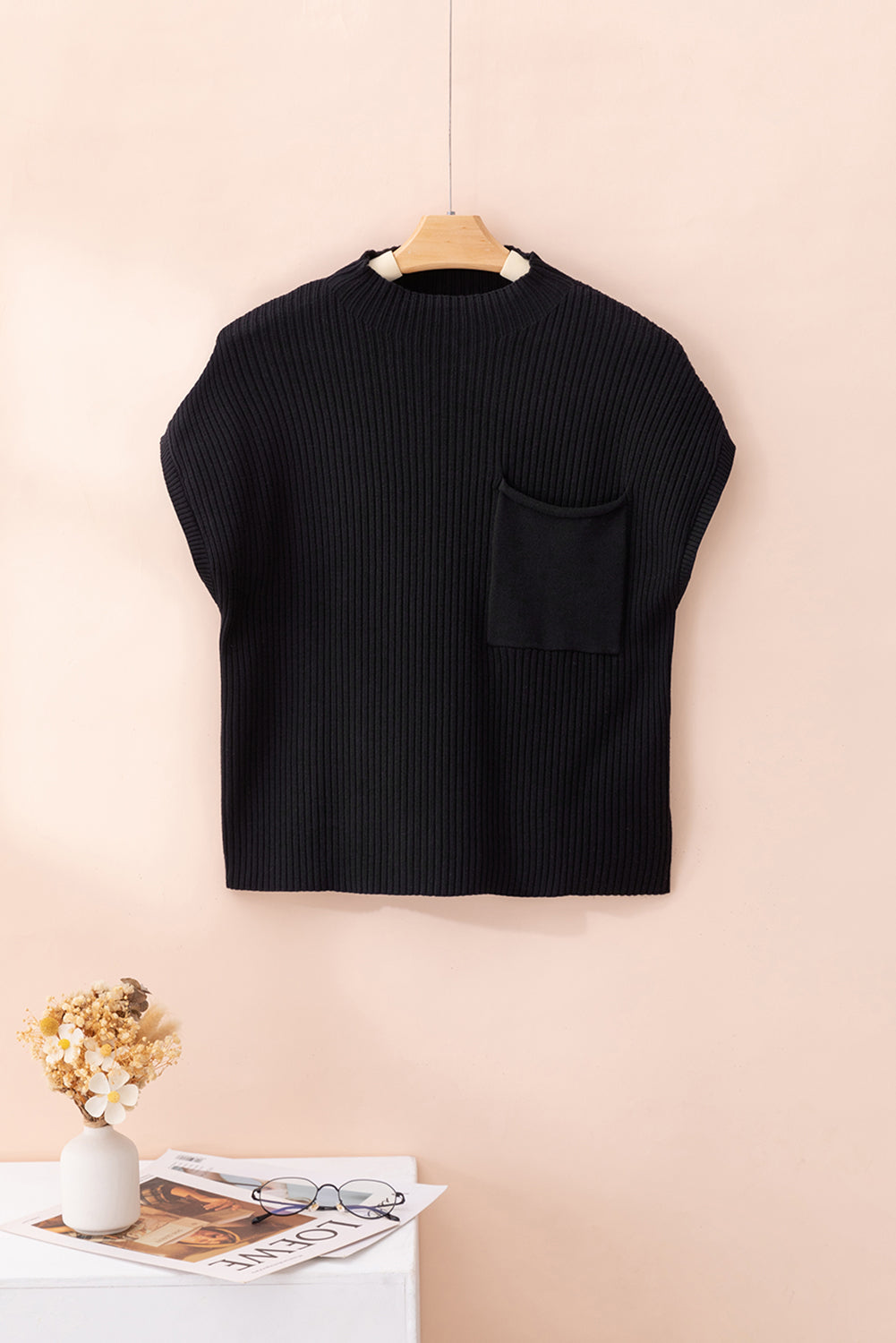 The Shewin Patch Pocket Ribbed Knit Short Sleeve Sweater, elegantly displayed on a wooden hanger against a peach background, combines style with versatility. Below it, a table holds flowers, a book, and glasses—perfect for transitional weather.