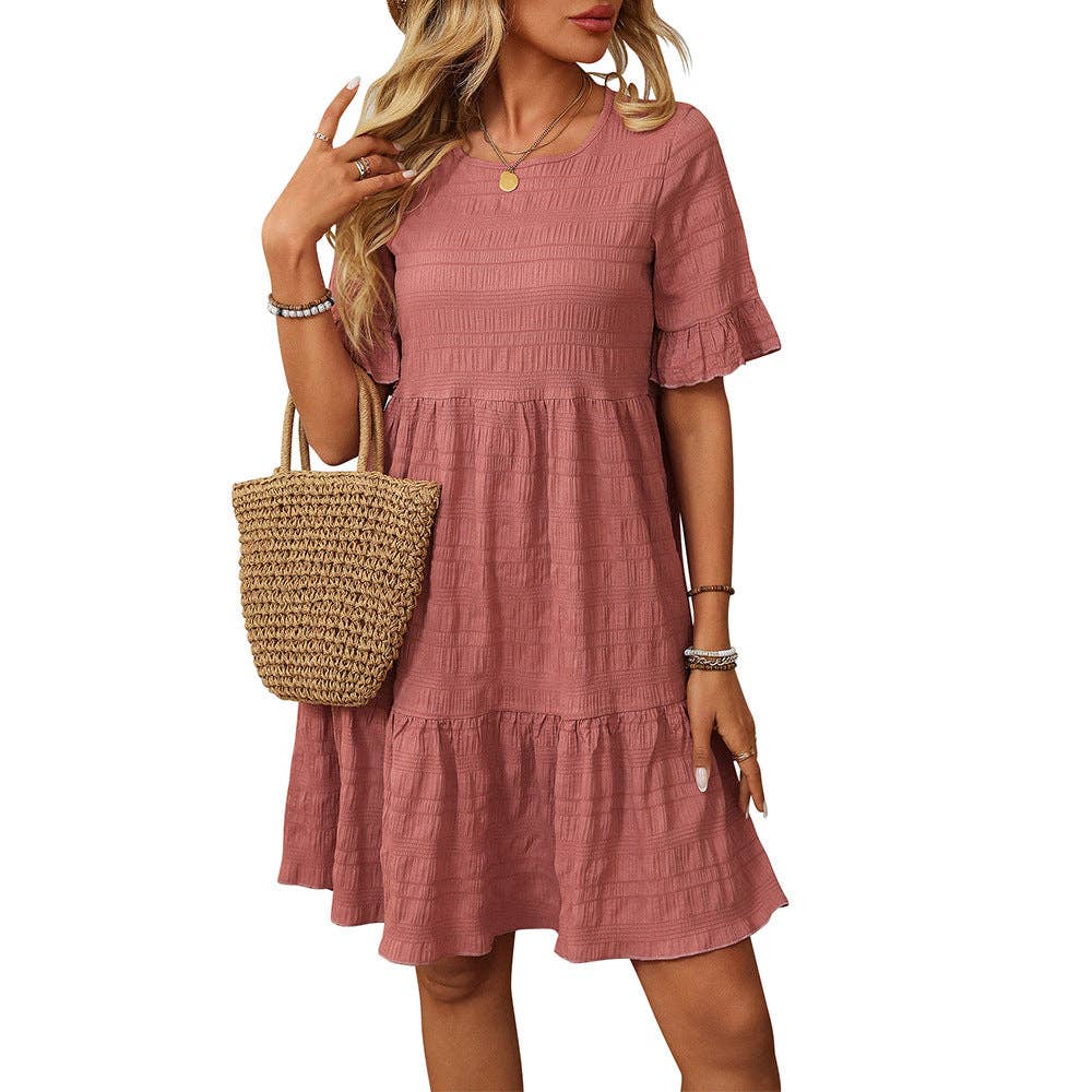 A woman wears the Suhkasana Loose Casual Short-Sleeve Dress in textured pink, paired with a woven straw handbag and accessorized with bracelets and a necklace.