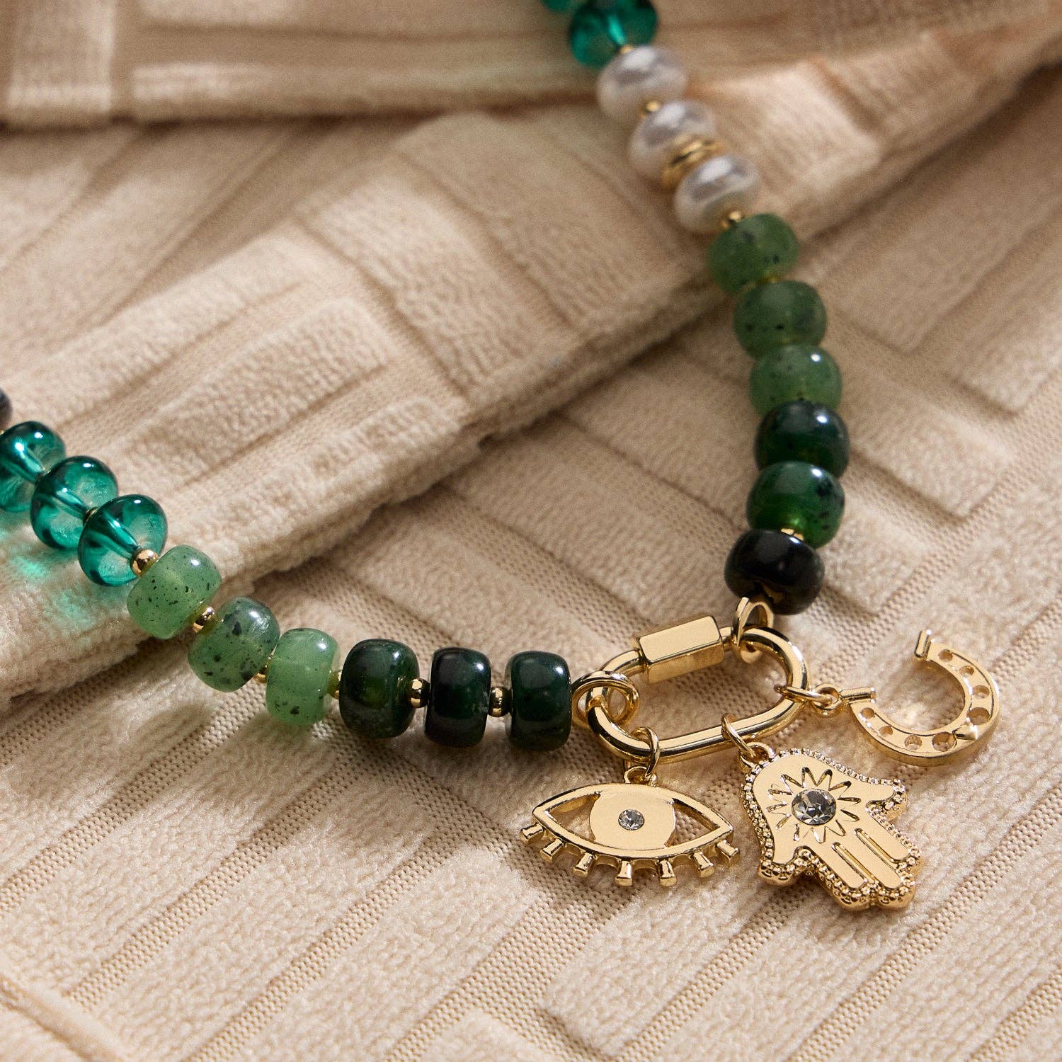 The Lilla Haven Textured Glass Bead Necklace with Charm Trio features green beaded glass, gold-toned spacers, and three gold charms—an eye, a hamsa hand, and a horseshoe—displayed on beige textured fabric for an elegant, layered accessory look.
