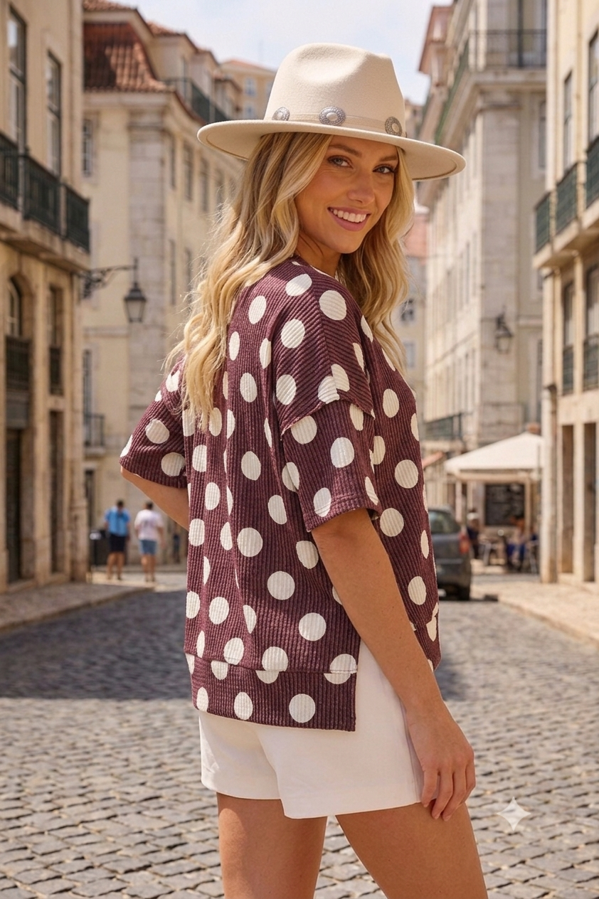 A woman in a beige hat, white shorts, and the Celeste Clothing Women’s Polka Dot Rib Knit Top stands on a cobblestone street lined with buildings, smiling at the camera.