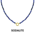 A Semi Precious Natural Stone Charm Keeper Necklace by WJW, featuring round blue sodalite beads and a central 18K Gold PVD Coated clasp, is displayed against a white background. Below the necklace, the word "SODALITE" appears.