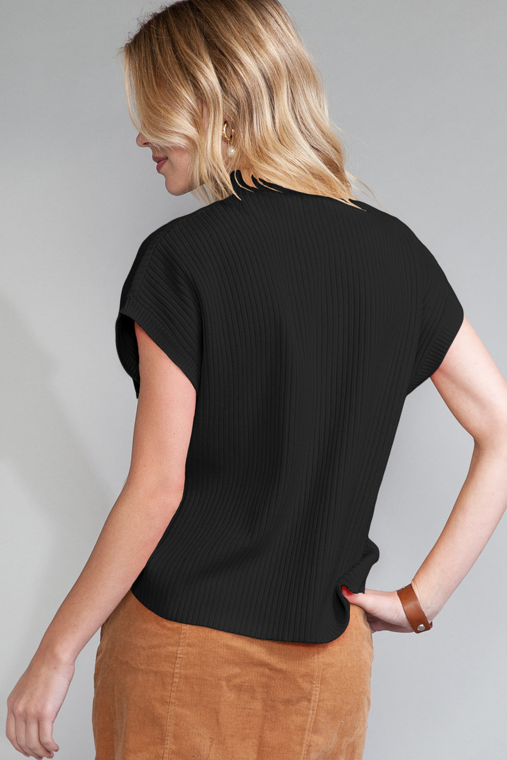 A blonde person dons a Shewin Patch Pocket Ribbed Knit Short Sleeve Sweater in black and tan pants, turned slightly to the side against a gray background.