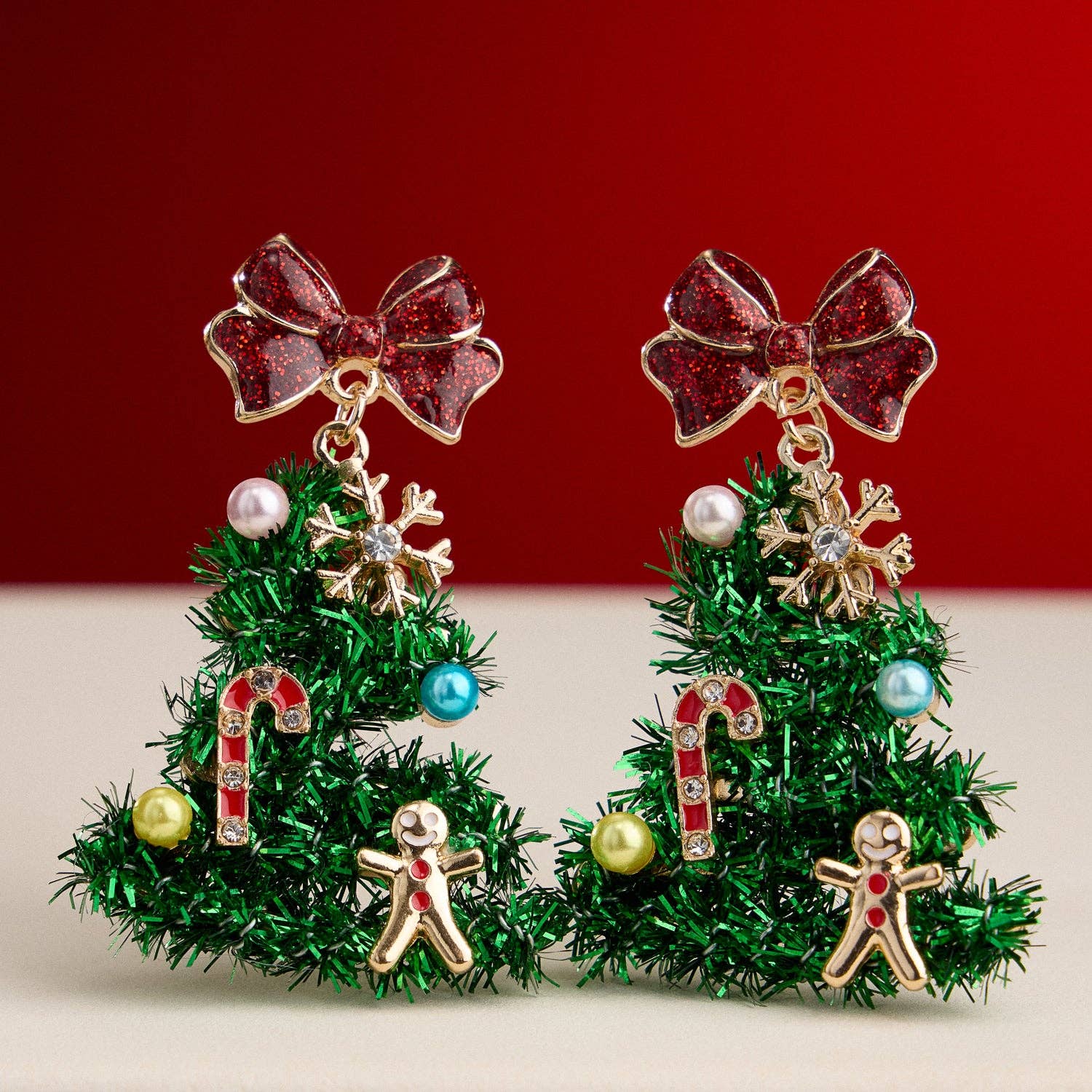 Adorn your holiday look with the Tinsel Christmas Tree Drop Earrings by Lilla Haven—festive ornament charm earrings shaped like Christmas trees and beautifully embellished with red bows, candy canes, gingerbread men, snowflakes, and colorful beads. Set against a cheerful red and beige backdrop, these earrings are the perfect sparkling accessory for any festive ensemble.