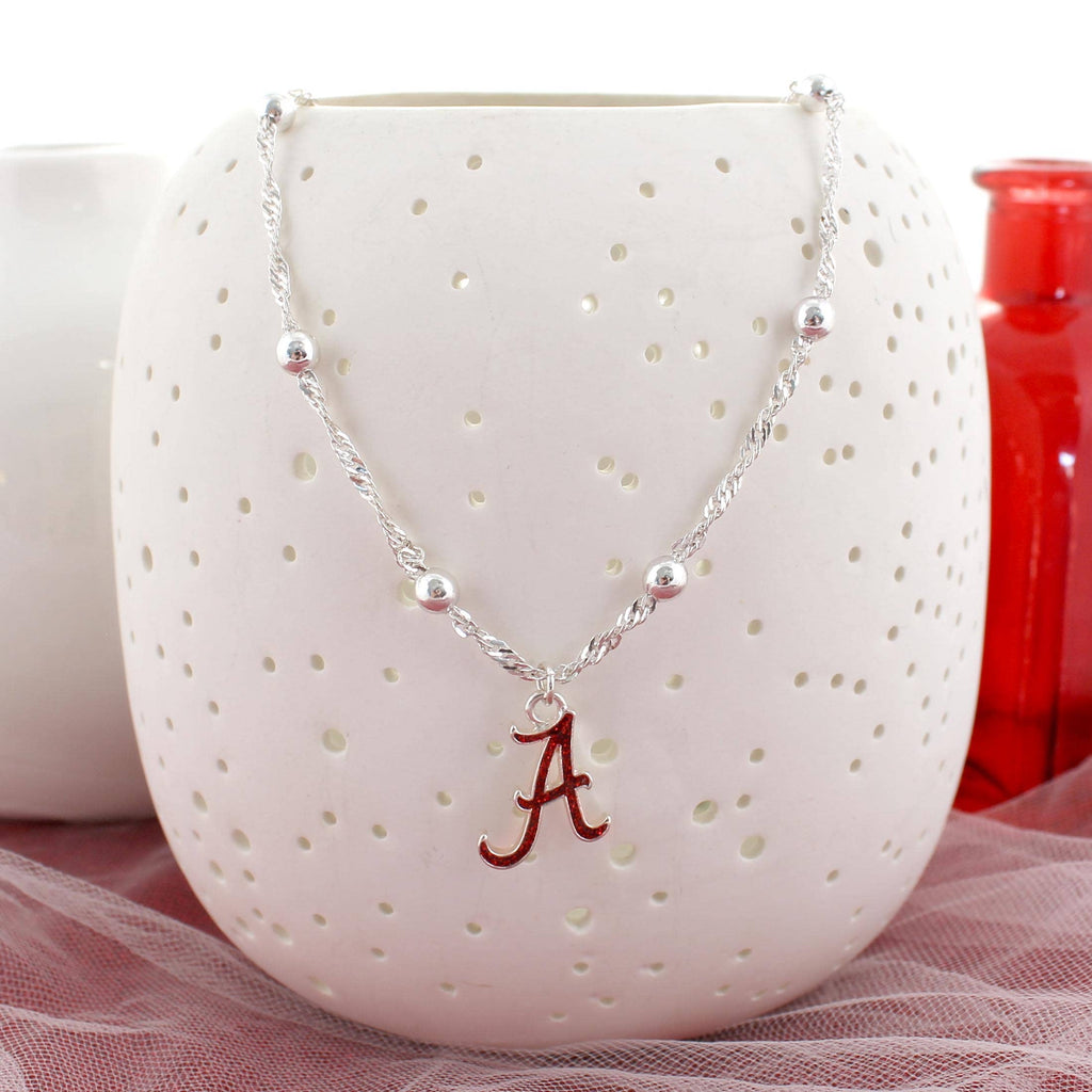 The Alabama Glitter Enamel Logo Necklace by Seasons Jewelry, featuring a silver beaded chain and a sparkling crimson "A" pendant, hangs on a white perforated vase. Red and white containers in the background enhance the necklace’s vibrant University of Alabama-themed display.