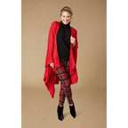 A woman wearing the COCO + CARMEN Kiara Lightweight Wrap over a black turtleneck, red plaid pants, and black boots stands against a plain beige background.