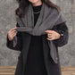 A woman wears the JC Sunny Fashion Cashmere-Blend Knit Triangle Shawl with a cross-front design over a dark coat, standing on a wooden floor against a textured gray wall.