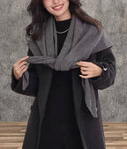 A woman wears the JC Sunny Fashion Cashmere-Blend Knit Triangle Shawl with a cross-front design over a dark coat, standing on a wooden floor against a textured gray wall.