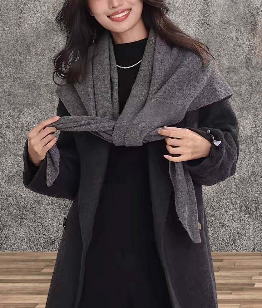 A woman wears the JC Sunny Fashion Cashmere-Blend Knit Triangle Shawl with a cross-front design over a dark coat, standing on a wooden floor against a textured gray wall.