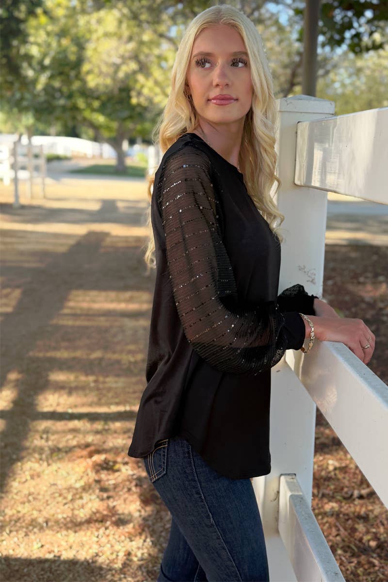 A woman with long blonde hair stands outdoors by a white fence, wearing the 24-Hrs Fashion Sequin Sleeve Blouse and blue jeans, with trees and sunlight in the background.