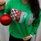 A person holds a Green Merry Sequin Christmas Sweatshirt with the word “merry” appliqued on the front in red, white, and green patterns, alongside a red disco-ball ornament.