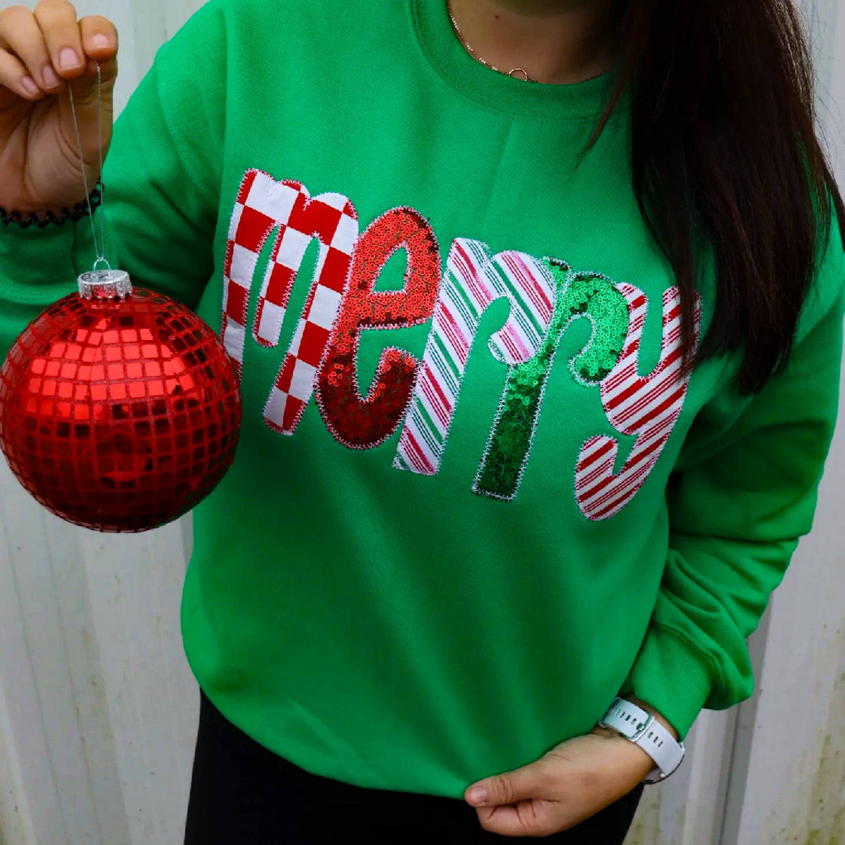 A person holds a Green Merry Sequin Christmas Sweatshirt with the word “merry” appliqued on the front in red, white, and green patterns, alongside a red disco-ball ornament.