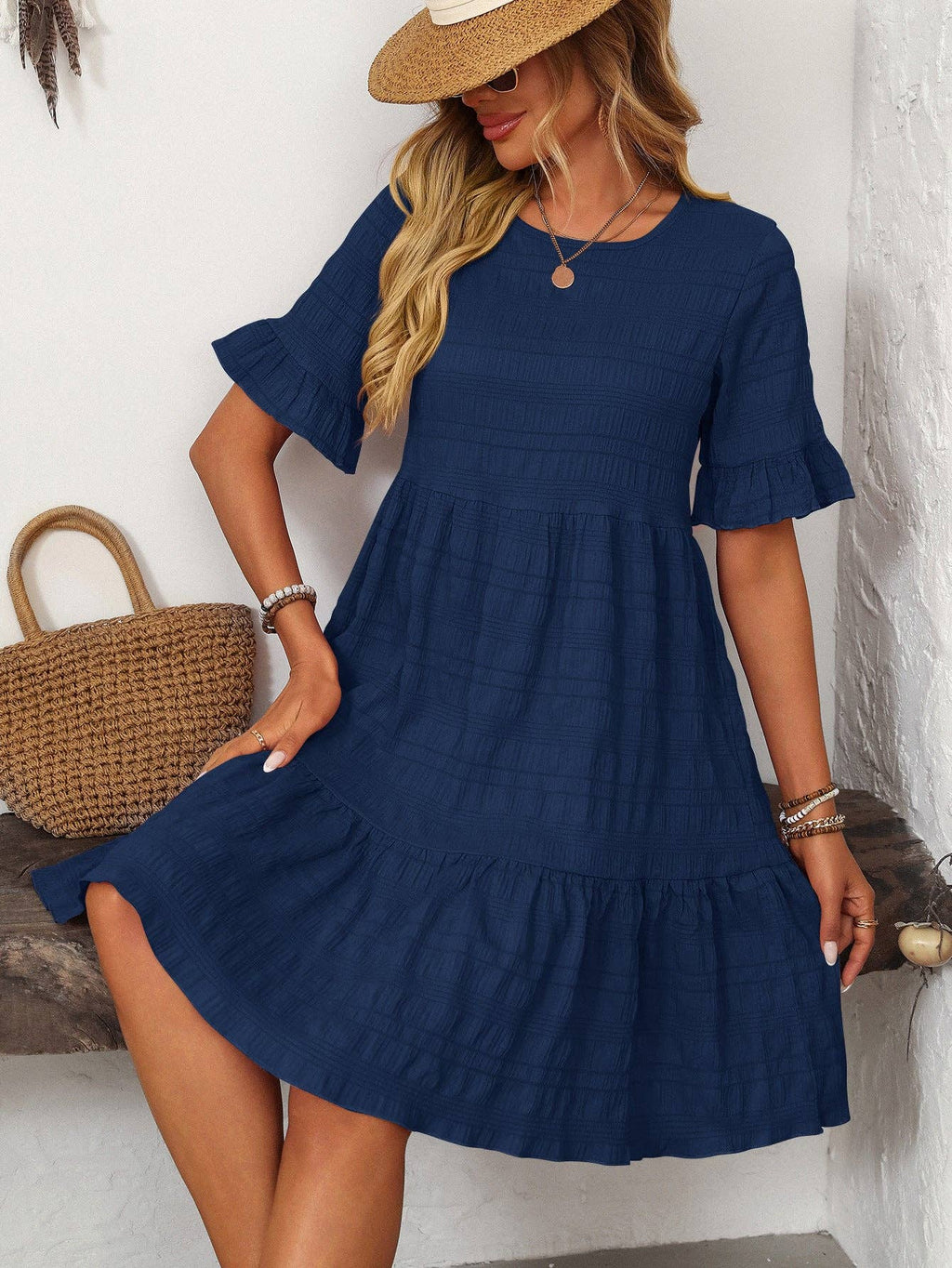 A woman in a Suhkasana Loose Casual Short-Sleeve Dress and straw hat sits on a bench beside a woven bag—ideal as an elegant summer outfit for relaxed days.